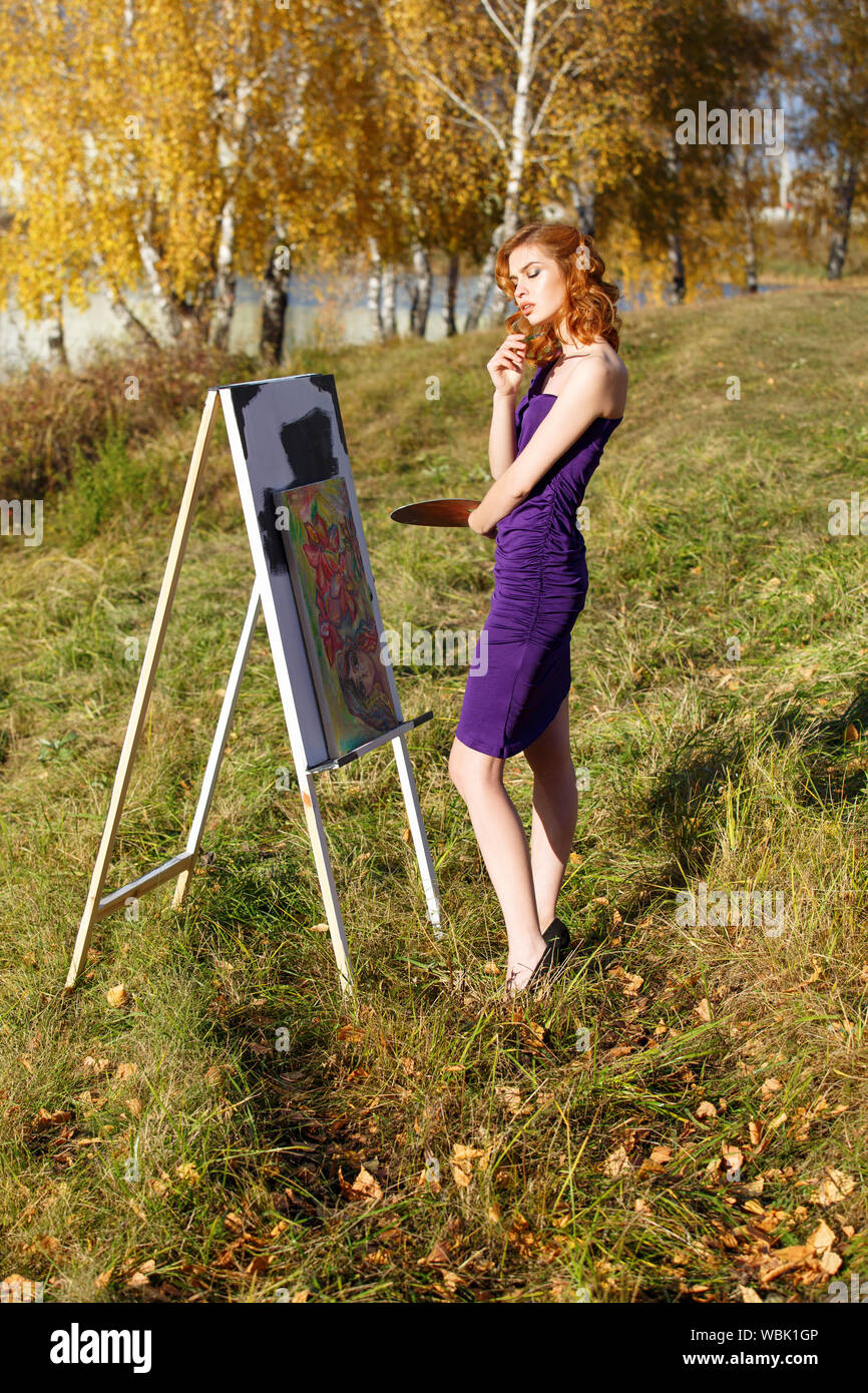 Young beautiful woman in sexy purple dress with palette and brush painting outdoors in autumn park. Stock Photo