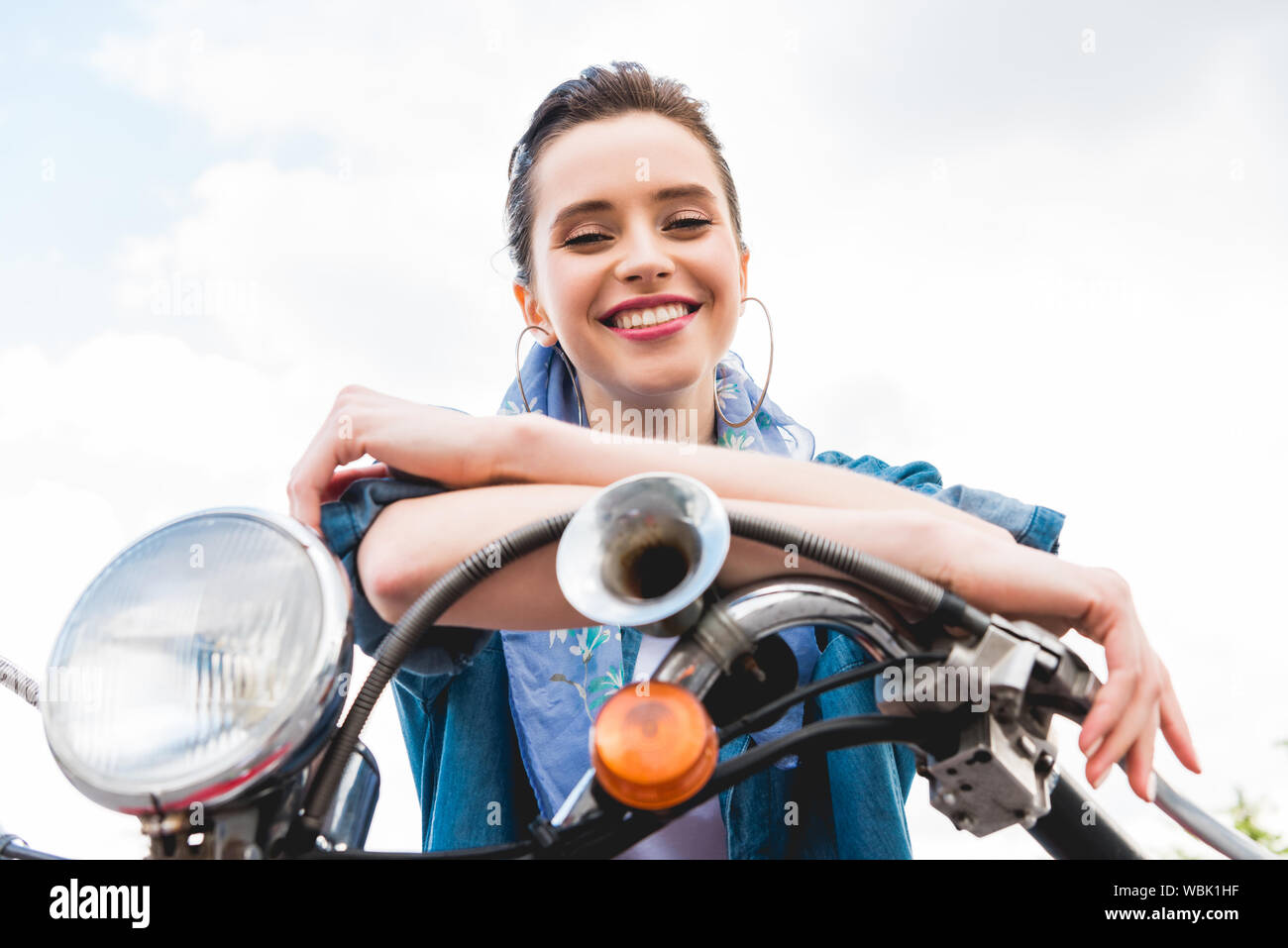 low angle view of beautiful girl sitting on scooter, resting, looking at camera and smiling on sky background Stock Photo