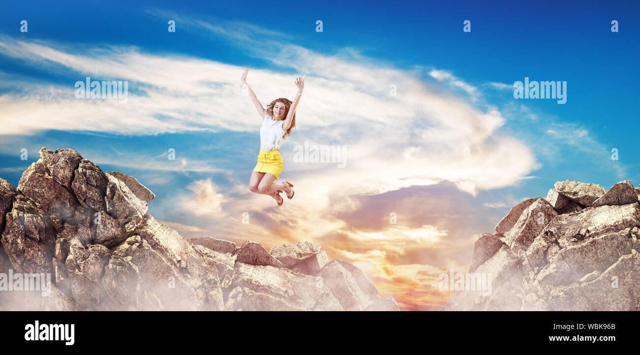 Happy business woman jumps up on the gap between hills over cliff on the blue sky background. Stock Photo