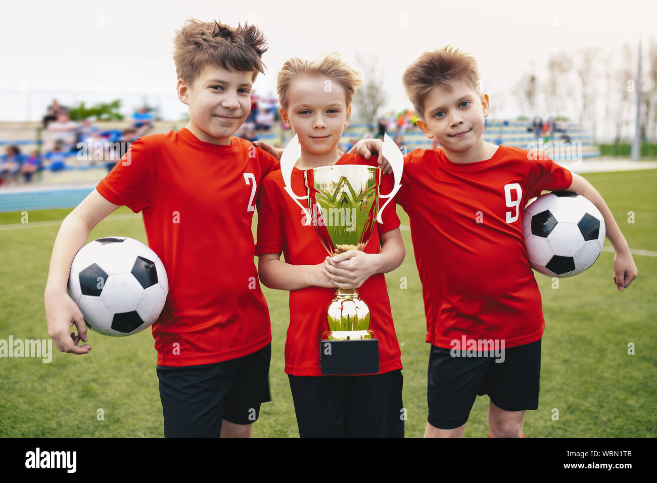 Three Happy Cheerful Kids of Sports Soccer Team. Boys Football Players Holding Trophy at the Stadium. Young Winners of Youth Football Tournament Stock Photo