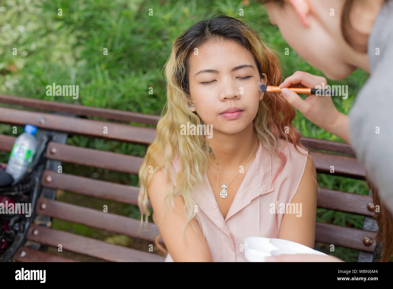 Female Asian model sitting down on a park bench while applying makeup to her cheeks Stock Photo