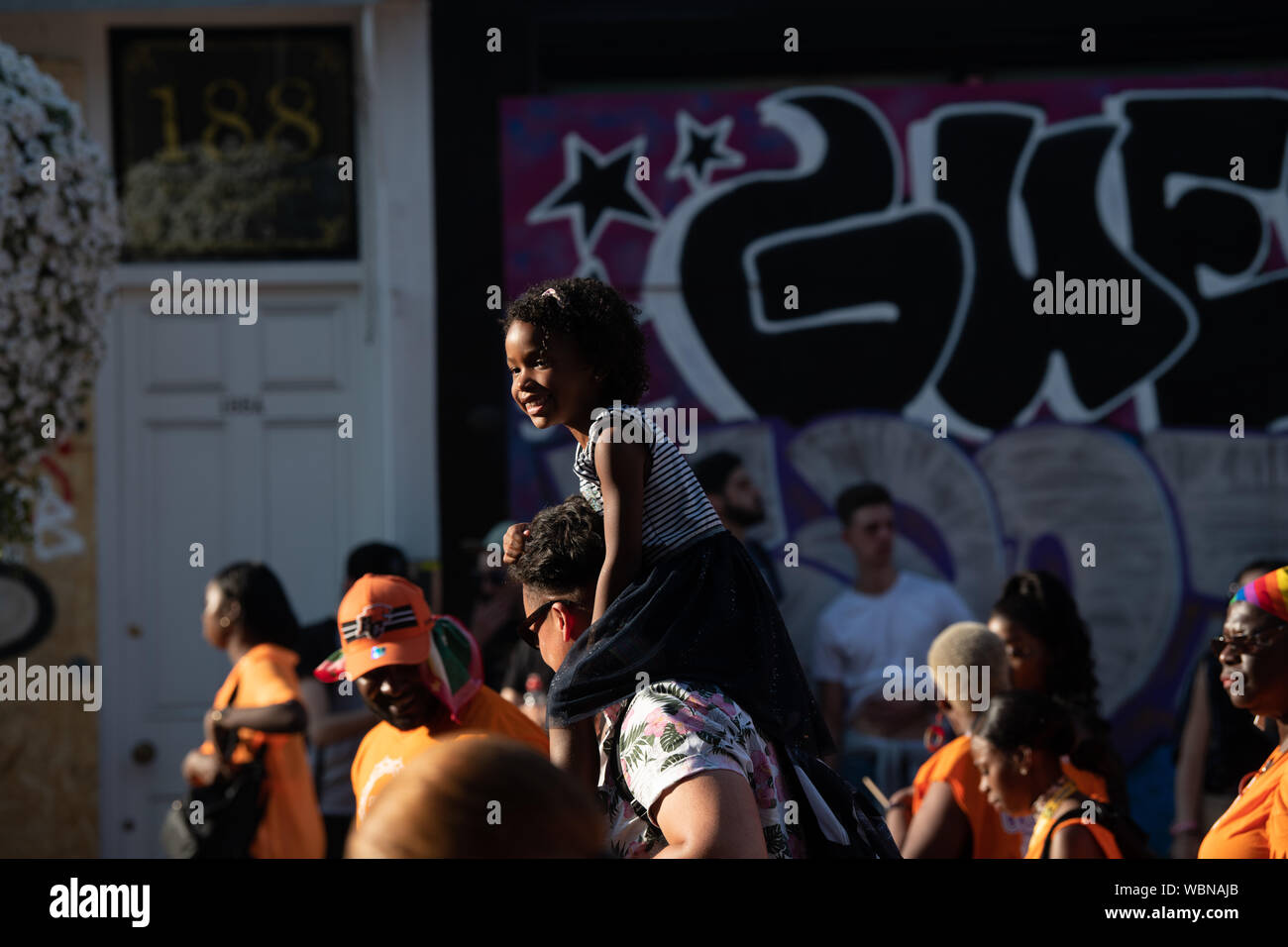 A smiling young black girl sitting on her father's shoulders whilst walking through a crowd at Notting Hill Carnival Stock Photo