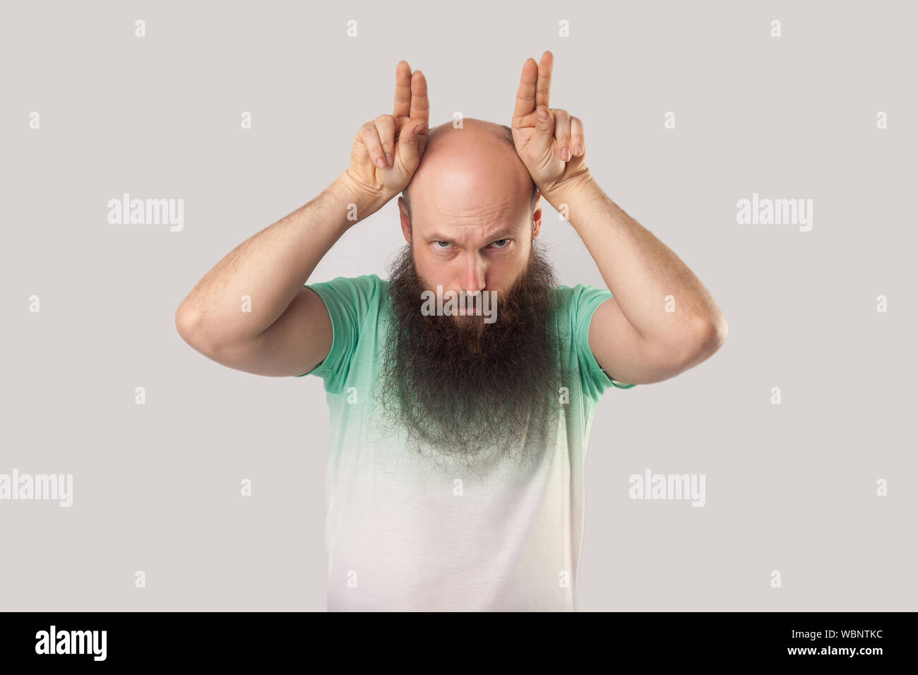 Portrait of serious or angry middle aged bald man with long beard in green t-shirt standing with finger horns on head and looking with aggressive face Stock Photo