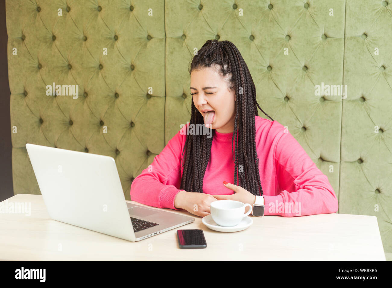 Portrait of beautiful childish funny young girl freelancer with black dreadlocks hairstyle in pink blouse sitting and showing tongue out on webcamera Stock Photo