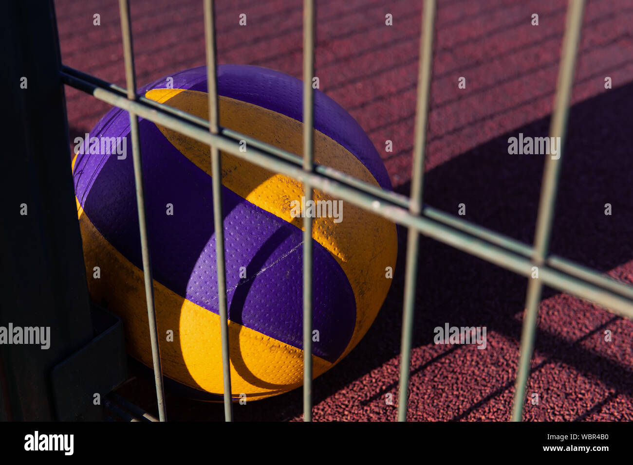 A lonely ball laying on the red ground after the game. Lost volleyball equipment. Sports field at the sunset with a metal fence. Stock Photo