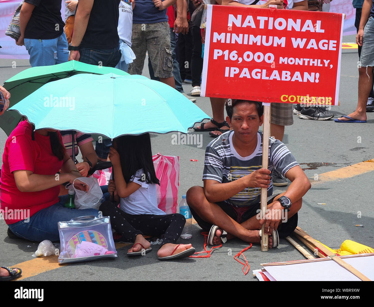A man holds a placard next to his family on the street, during the demonstration.Thousands of workers took to the streets as the Philippines marked National Heroes' Day. They call it the 'Martsa ng Manggagawa Laban sa Kontraktwalisasyon' (Workers' March Against Contractualization). They slammed the Duterte Government for allegedly turning a blind eye to the abuses suffered by workers. The United workers calling for an end to 'ENDO' or end of contractualization of workers and the increase of minimum wage of workers to 750 pesos. Stock Photo
