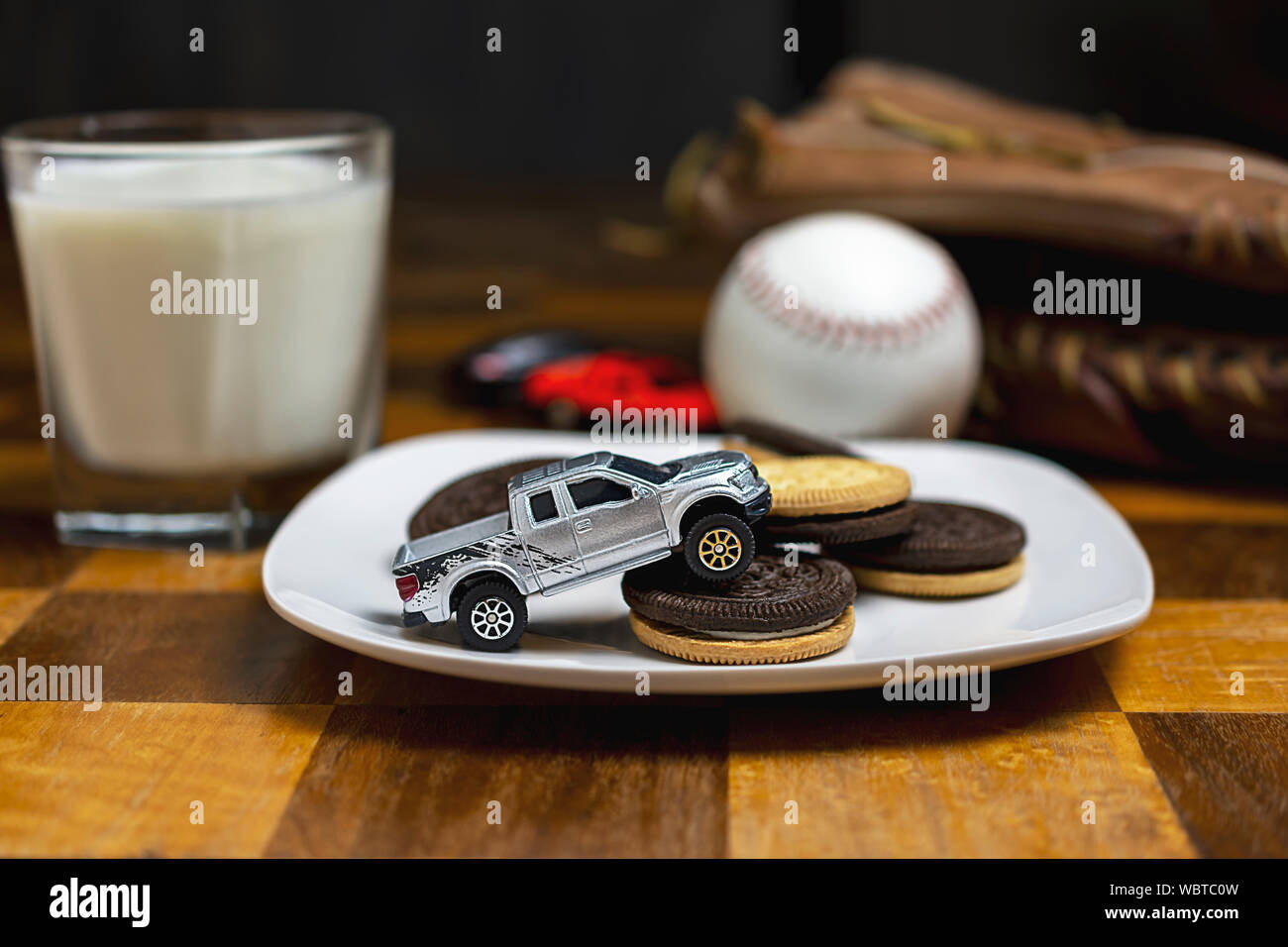 Toy truck driving over a plate of sandwich cookies.  Milk, ball and glove blurry in background. Stock Photo