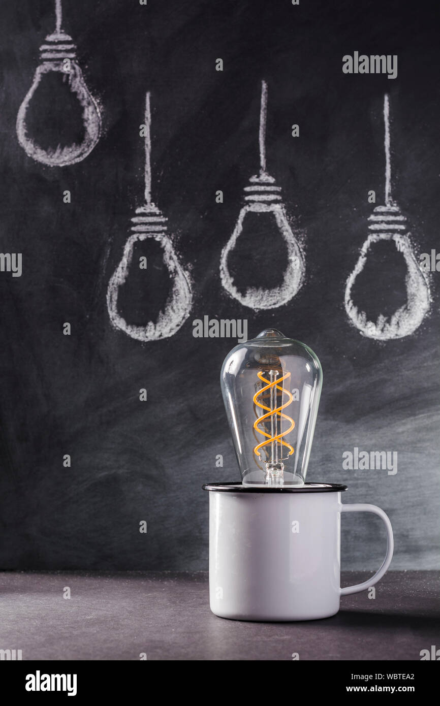 Light bulbs sketched in white chalk behind a tin mug holding and Edison bulb signifying growing ideas. Stock Photo