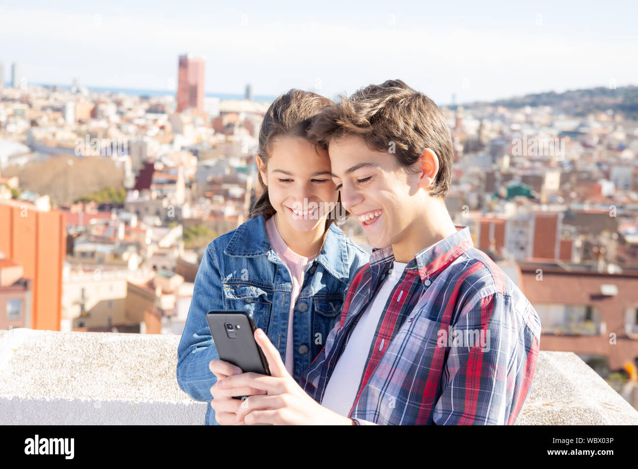 Dos niños sonrientes mirando el móvil en una terraza en el exterior Stock Photo
