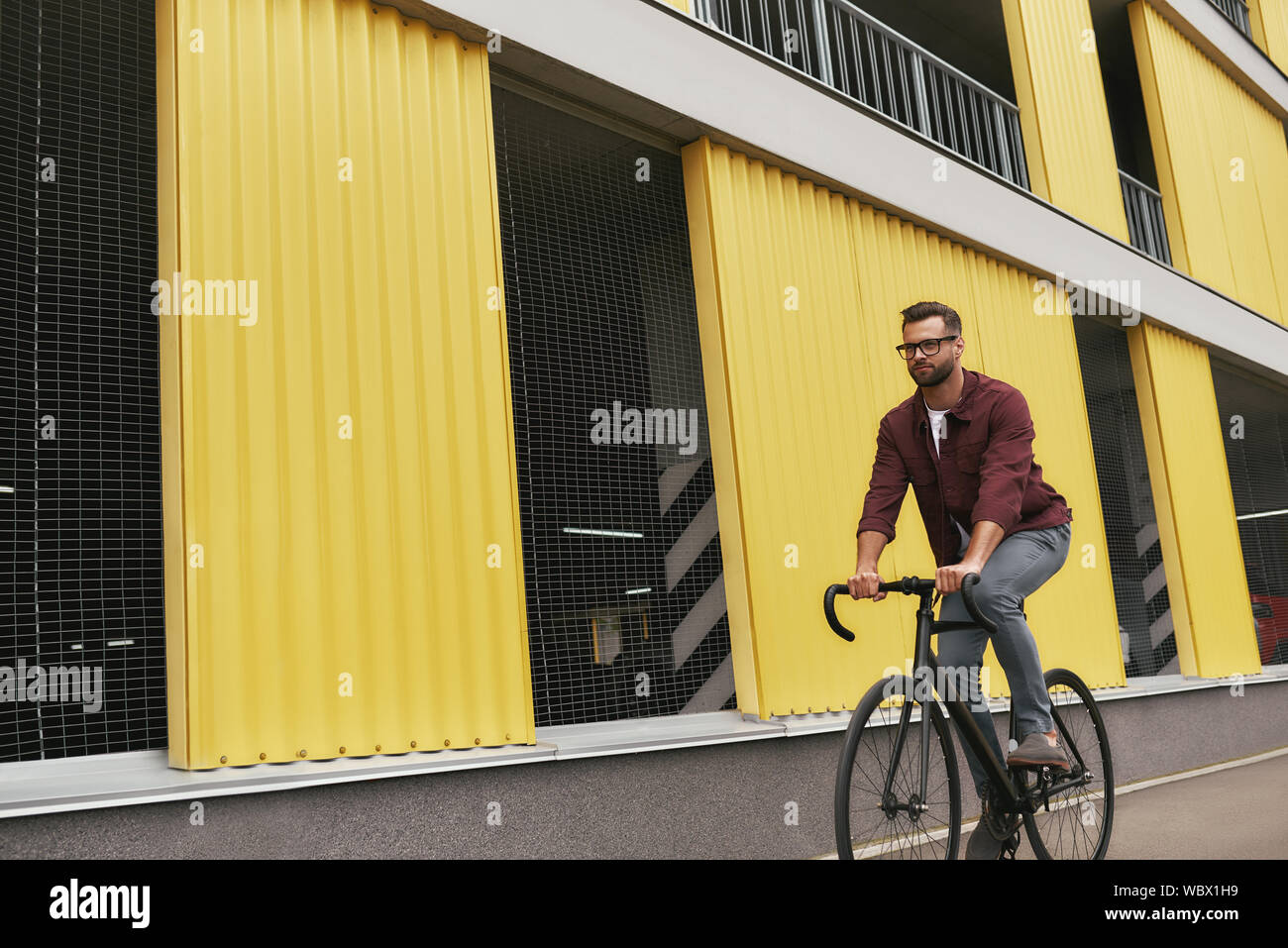 Free time. Handsome man with stubble in casual clothes riding on his bicycle along a road against yellow building. Weekend activity. Lifecycle Stock Photo