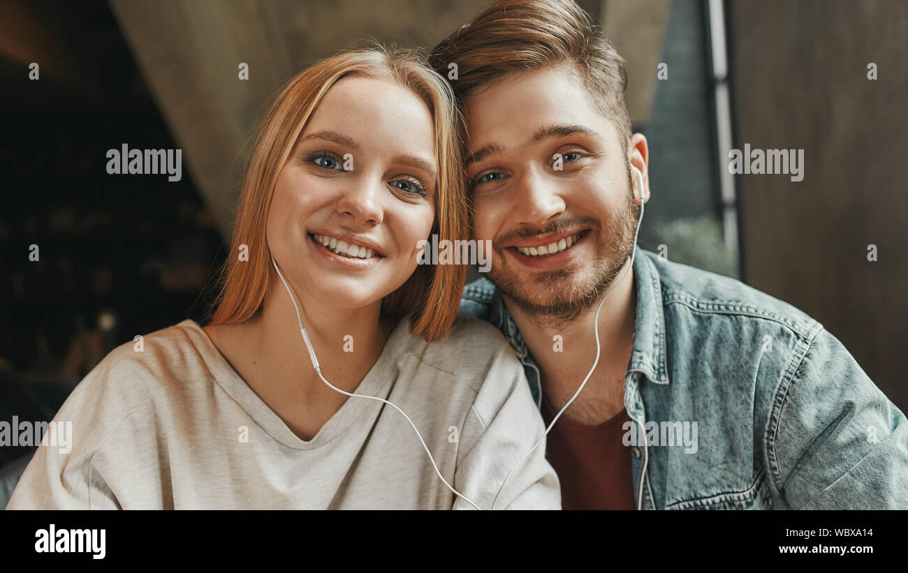 Portrait of caucasian man and woman looking at camera while listening to music via earphones together. They are sitting in a cafeteria during lunch. Horizontal shot. Front view Stock Photo