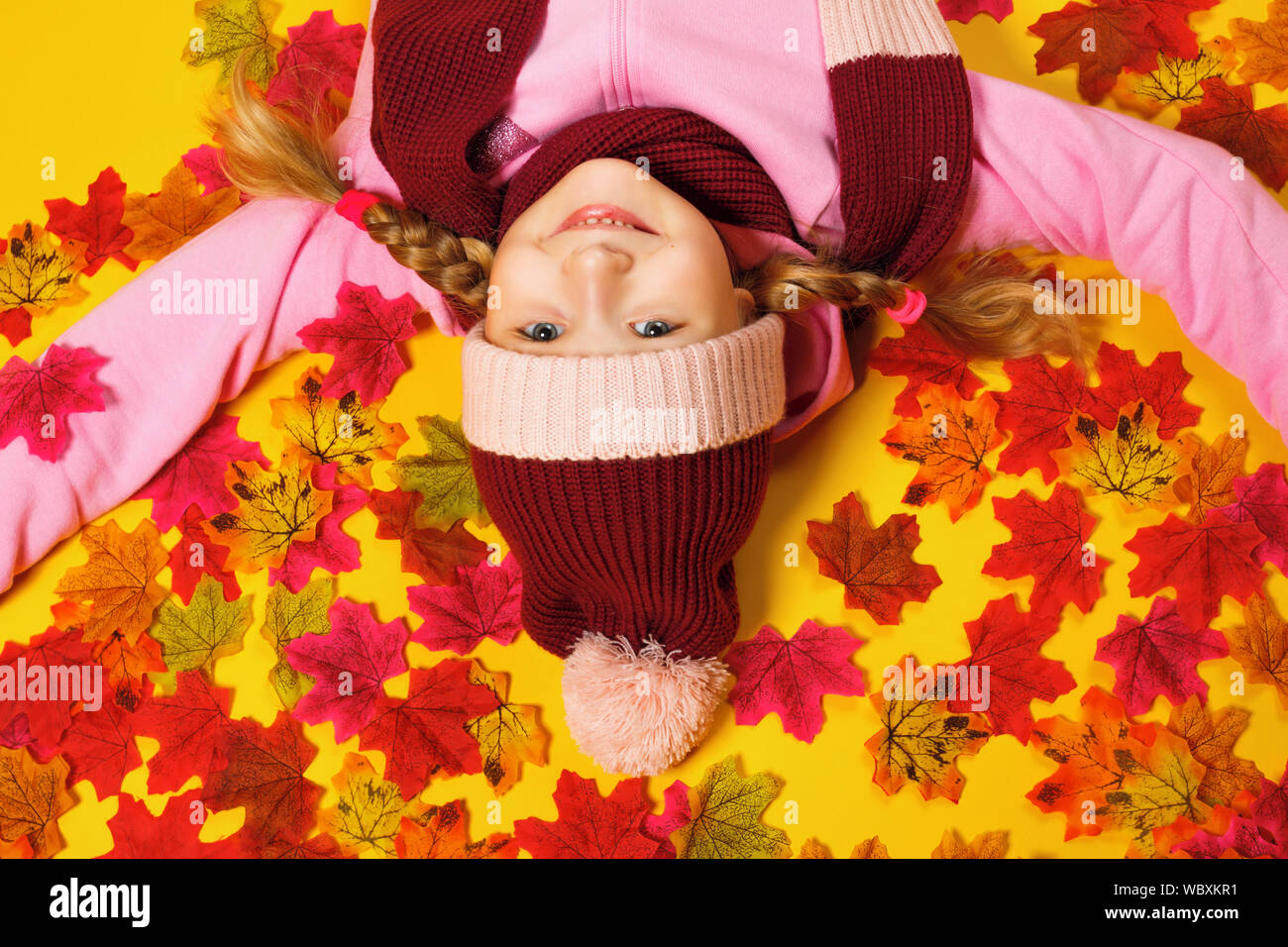 Top view of a happy little girl lying on autumn leaves. The child is upside down in a scarf and hat. Stock Photo
