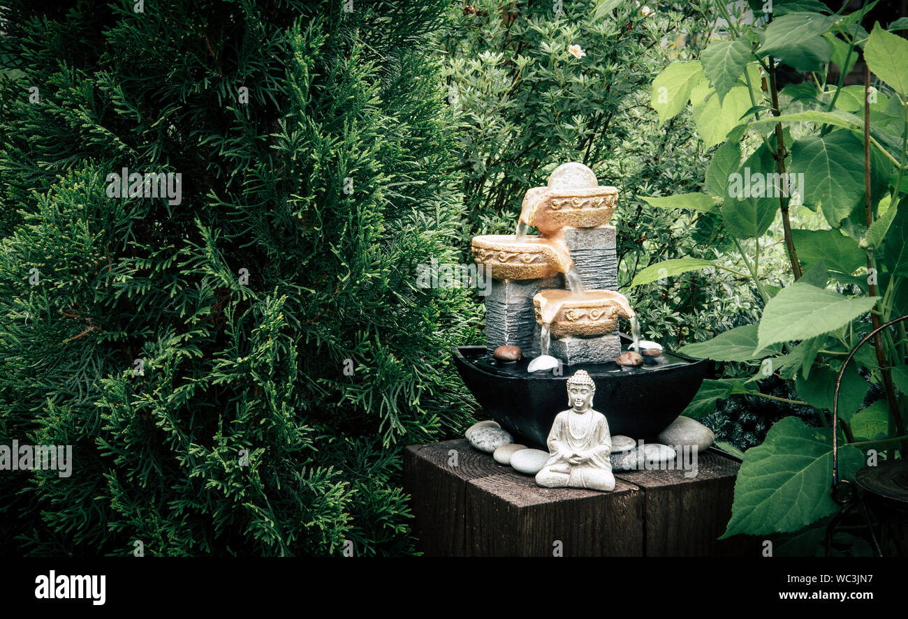 Small home garden private fountain with meditating Buddha statue between trees and bushes. Private zen garden concept. Stock Photo