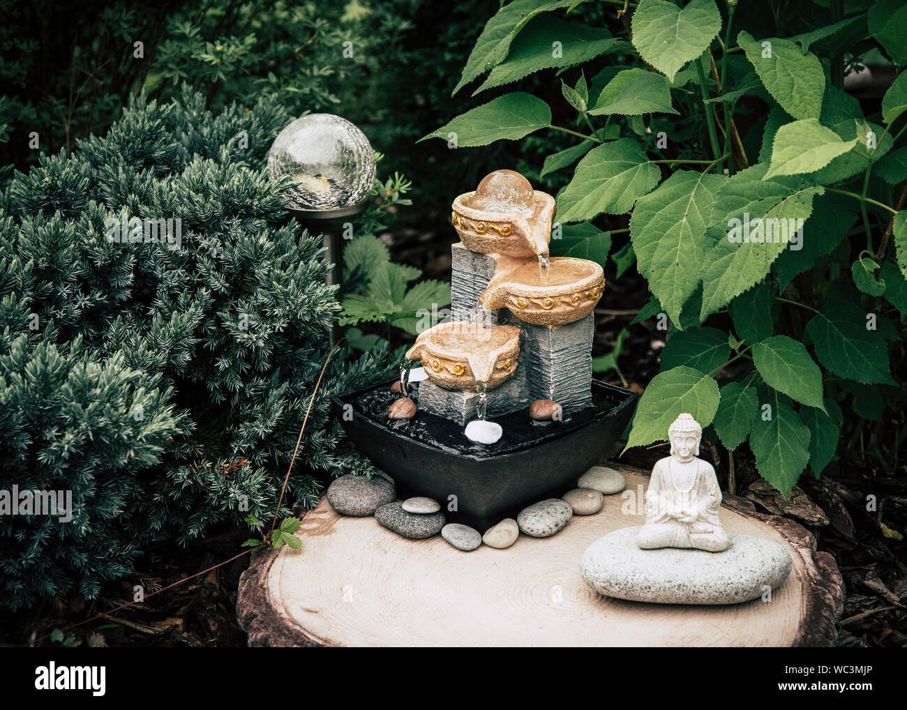 Small home garden private fountain with meditating Buddha statue between trees and bushes. Private zen garden concept. Stock Photo