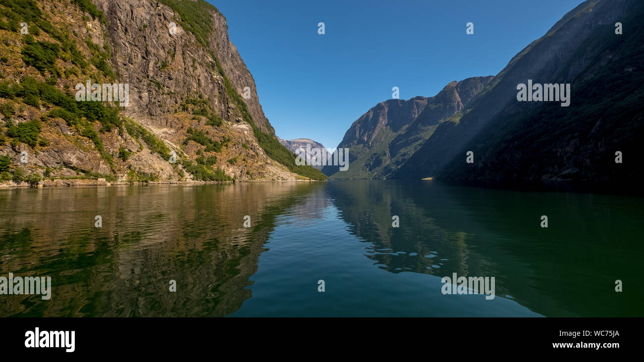 An approaching excursion ship on a beautiful fjord where the light is reflected, surrounded by rocks partly covered with trees, with a bright blue sky Stock Photo