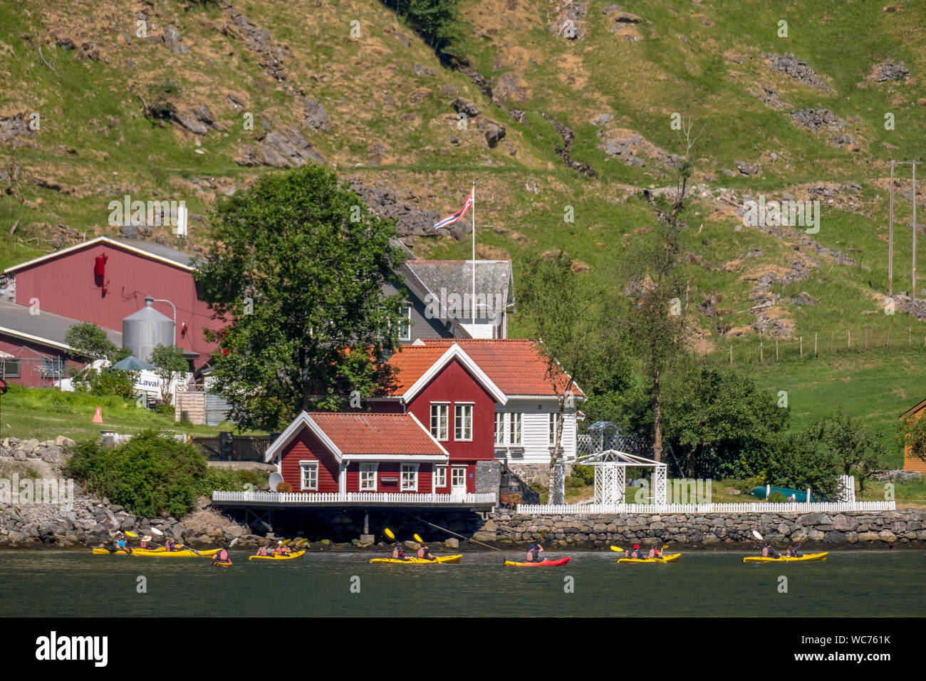 Tourists in yellow kayaks on Fjord, Flåm, Gudvangen, stone wall, mountain landscape, red wooden houses, Bakka, Sogn og Fjordane, Norway, Scandinavia, Stock Photo