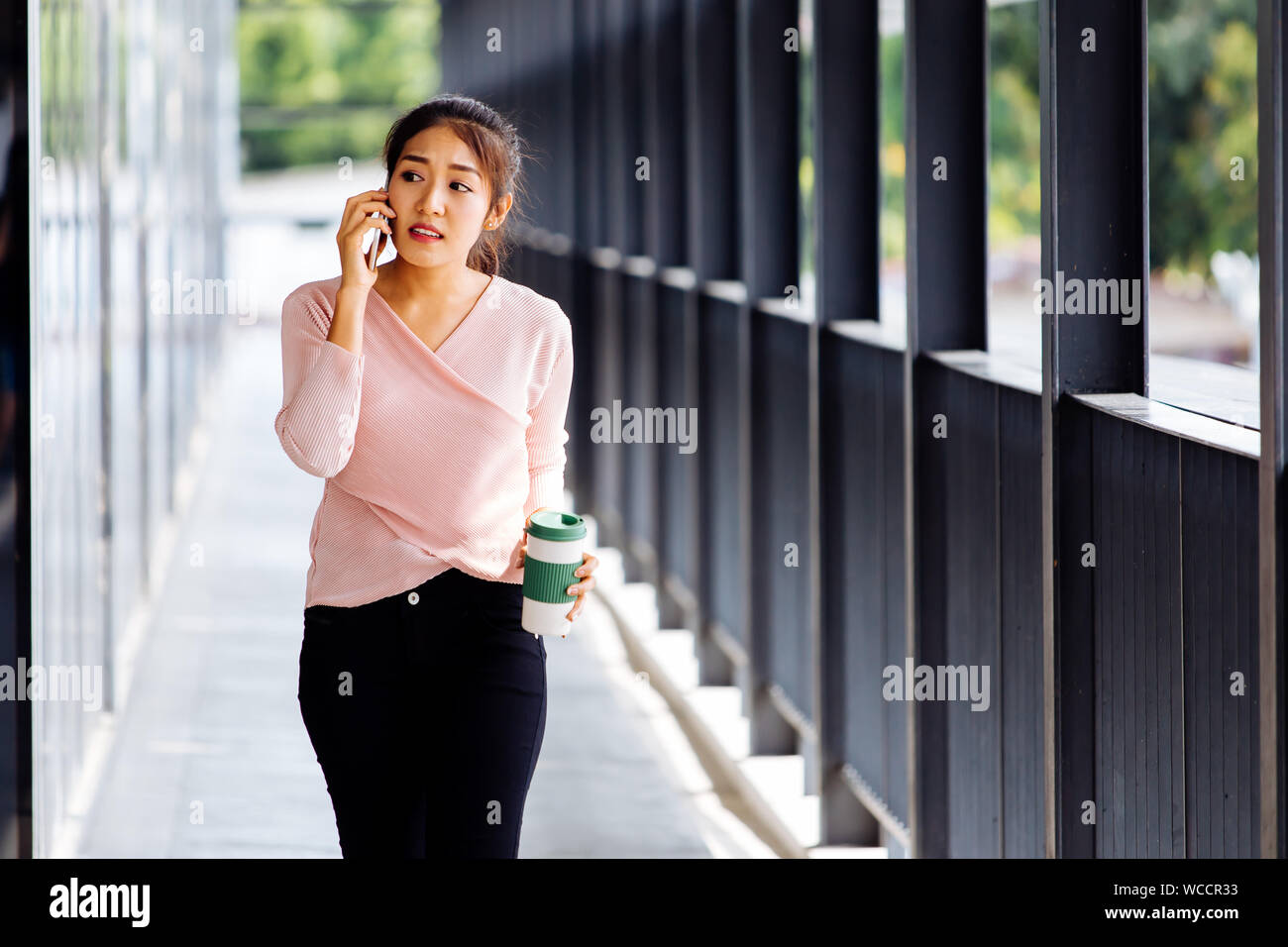 Attractive young fearful Asian lady drinking coffee on walk and speaking on mobile phone in outside corridor Stock Photo