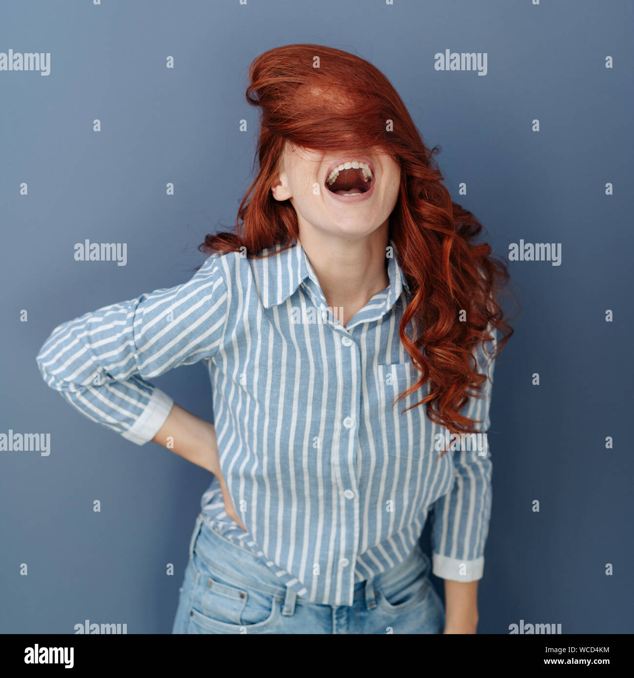 Young woman enjoying a good laugh as her long red hair flicks over her face  obscuring her eyes against a blue studio background Stock Photo - Alamy