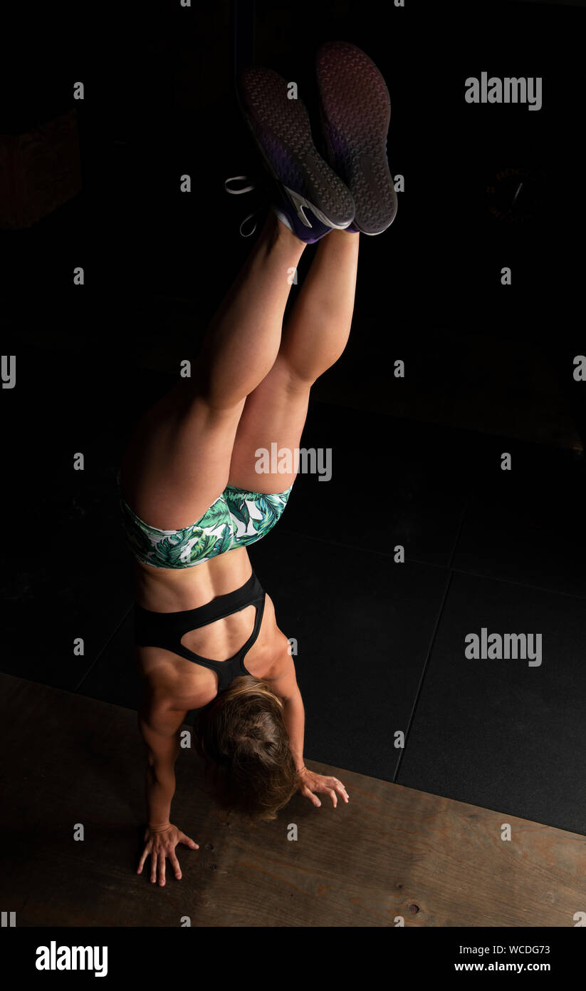 An attractive young woman with long hair and short pants is doing a handstand walk. Back muscles of a fit female athlete. Fitness workout in a gym. Stock Photo