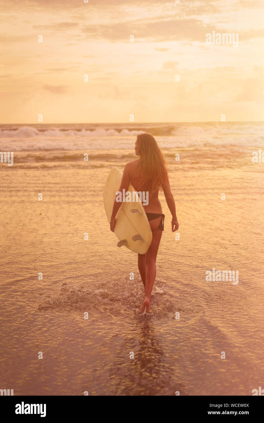 A young woman in bikini stands with her back to the camera and holds a surfboard by the ocean. Warm tinting. Surf concept. Full-length photo Stock Photo