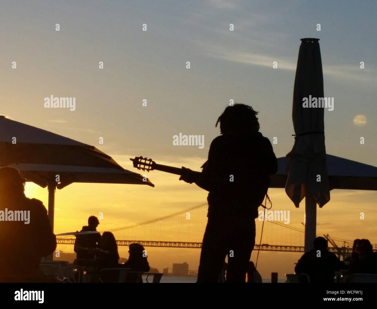 Silhouette Guitarist Playing Guitar At Cafe By Bridge Against Orange Sky Stock Photo
