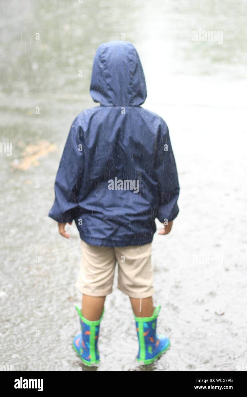 Rear View Of Boy In Raincoat Standing On Street During Rainfall Stock Photo