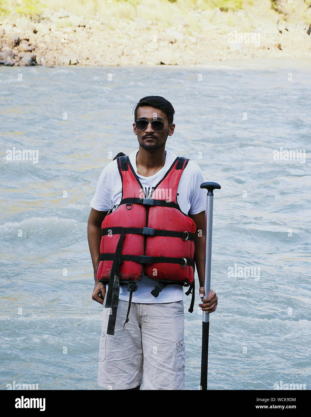 Young Man Wearing Life Jacket While Standing Against River Stock Photo