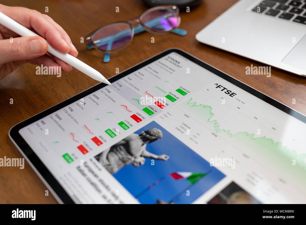 BATH, UK - AUGUST 28, 2019 : An Apple iPad Pro on a desk displaying stock market information using the Apple Stock app. A hand holding an Apple Pencil Stock Photo