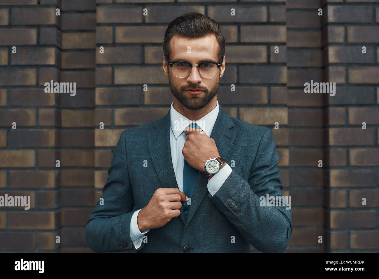 Perfect look. Confident businessman in full suit adjusting his tie and looking at camera while standing against brick wall. Business concept. Stylish people Stock Photo