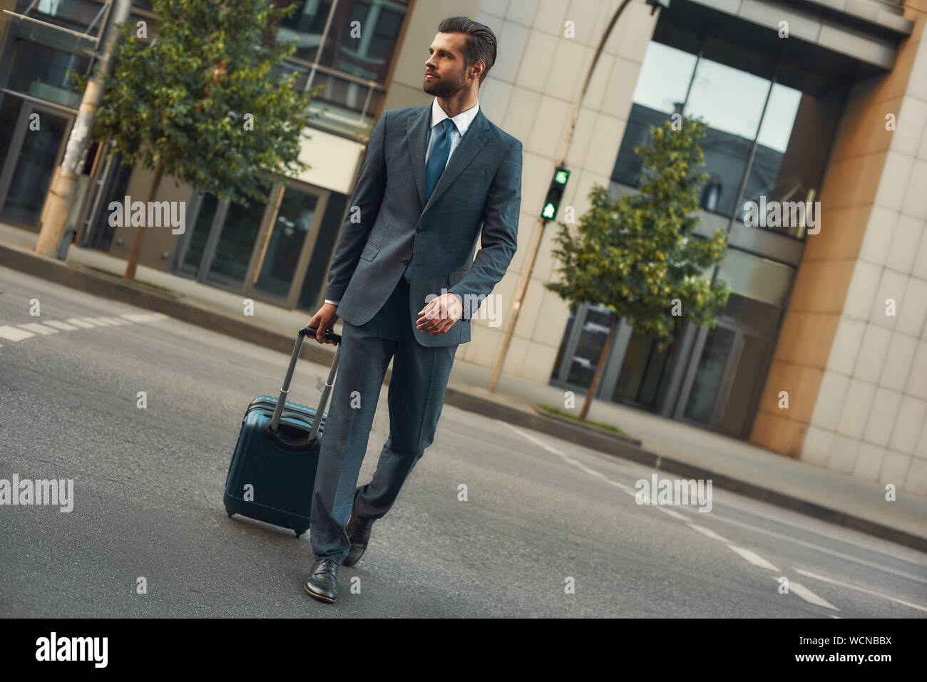 Business trip. Full length of young and handsome bearded man in suit pulling  his luggage and looking away while walking outdoors. Travelling Stock Photo  - Alamy