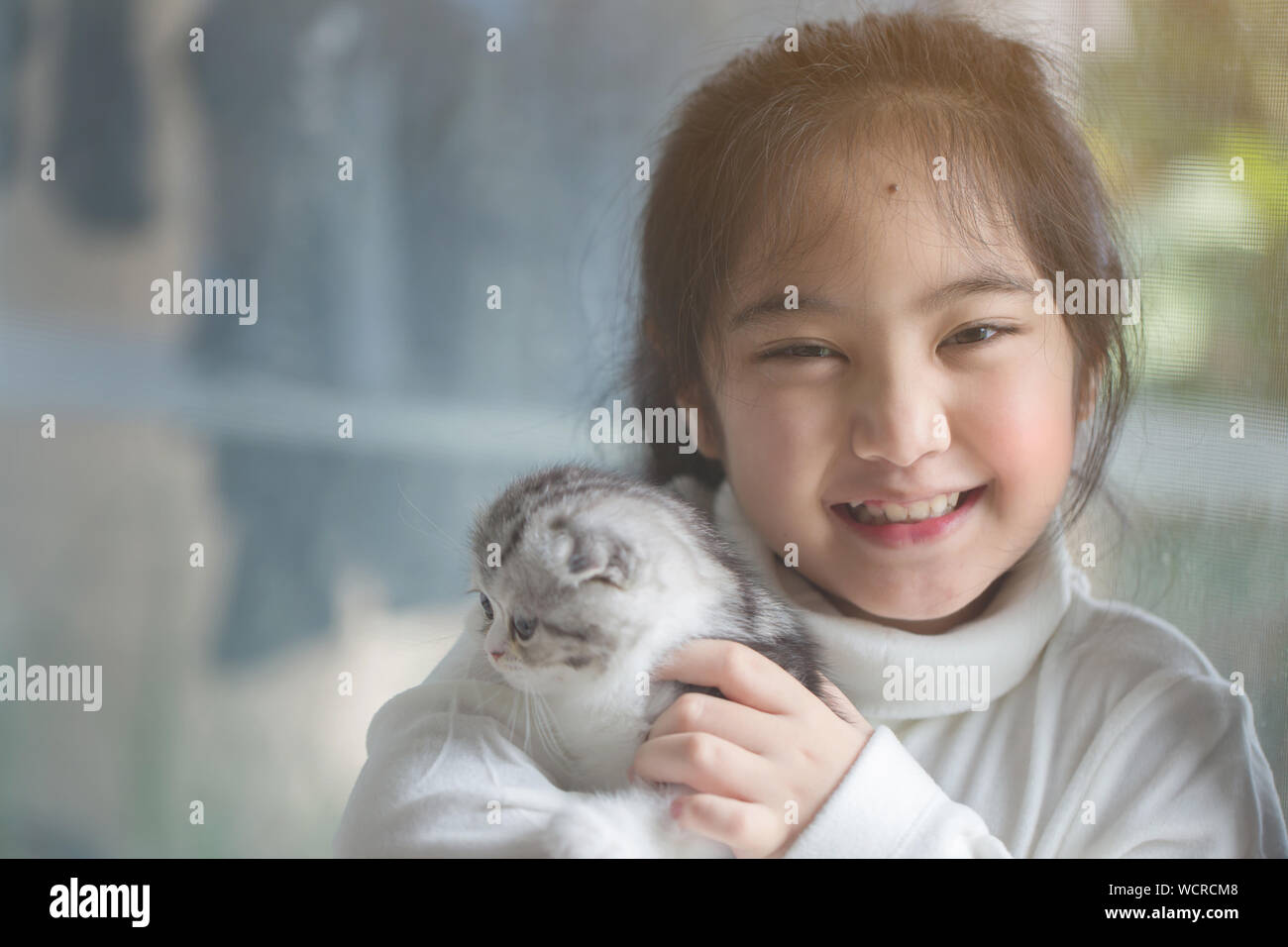 Portrait Of Happy Girl With Scottish Fold Cat Sitting By Window At Home Stock Photo
