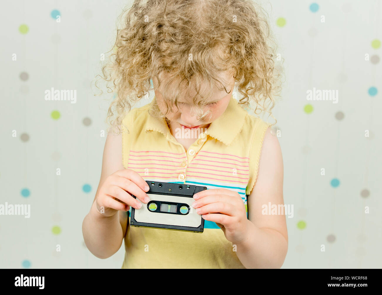 Funny picture of 5 year old girl holding and looking retro 80s cassette tape, being confused, what it is. Indoors, light spotty background. Stock Photo