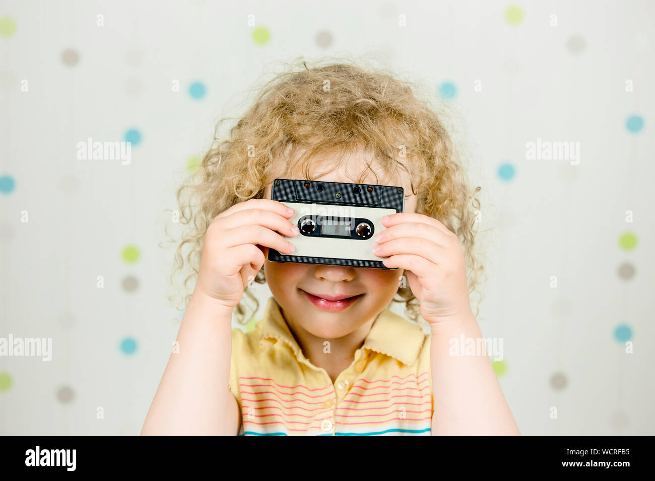 Funny picture of 5 year old girl holding and looking through retro 80s cassette tape indoors, light background. Stock Photo
