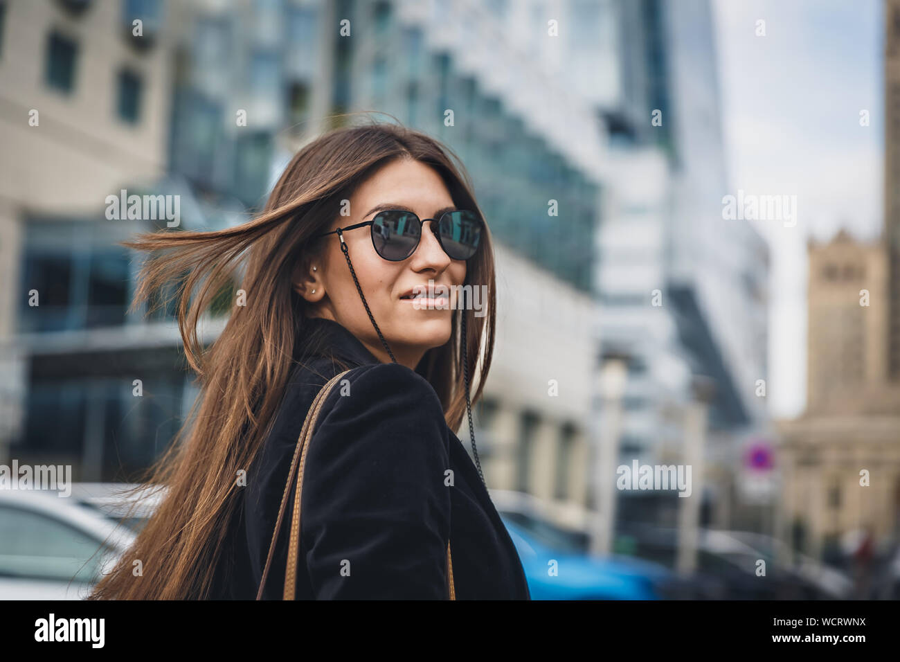 Young beautiful stylish girl in Sunglasses walking on street. Close up portrait of woman is turn around at camera and smiles. Female playfully looks Stock Photo