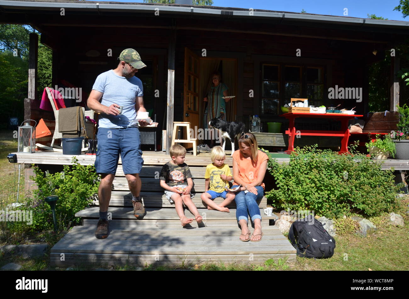 Two small boys and parents sitting in the sun outside a log cabin summerhouse near Ristiina, Finland. Stock Photo