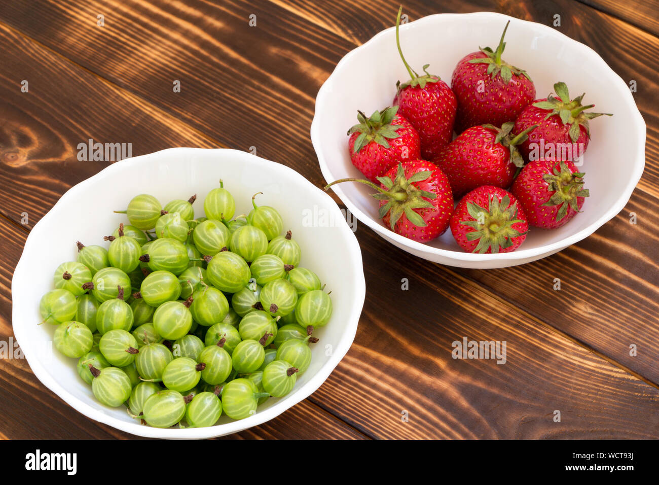 Green gooseberries and red ripened strawberries in white bowls on wooden table. Top view. Close-up. Natural organic homegrown products concept. Raw fo Stock Photo