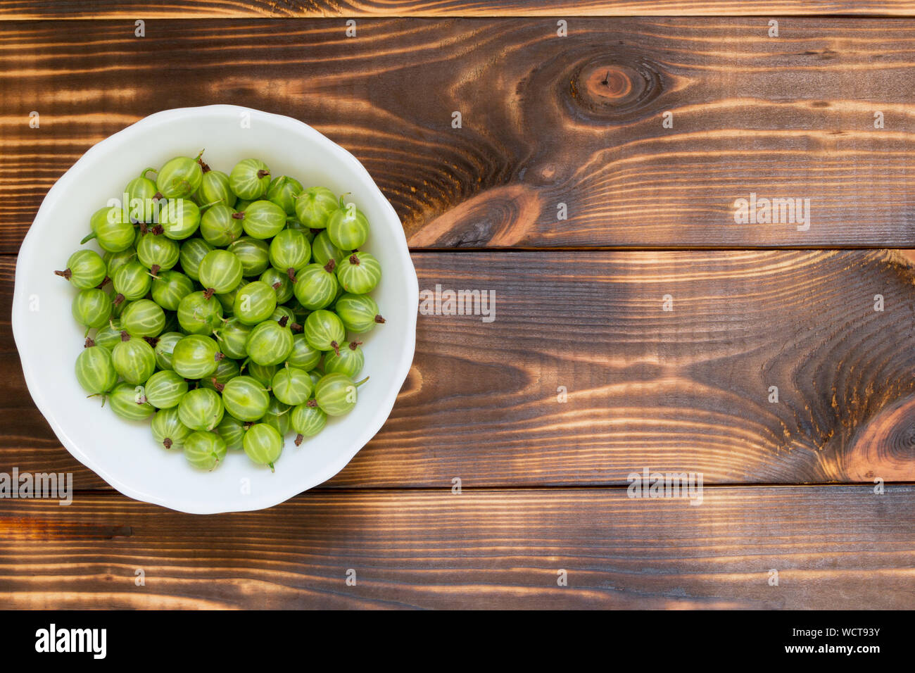 Green gooseberries on dark wooden table with copy space. Flat lay. Berries in white bowl at left side of image. Snack for raw diet. Natural homegrown Stock Photo