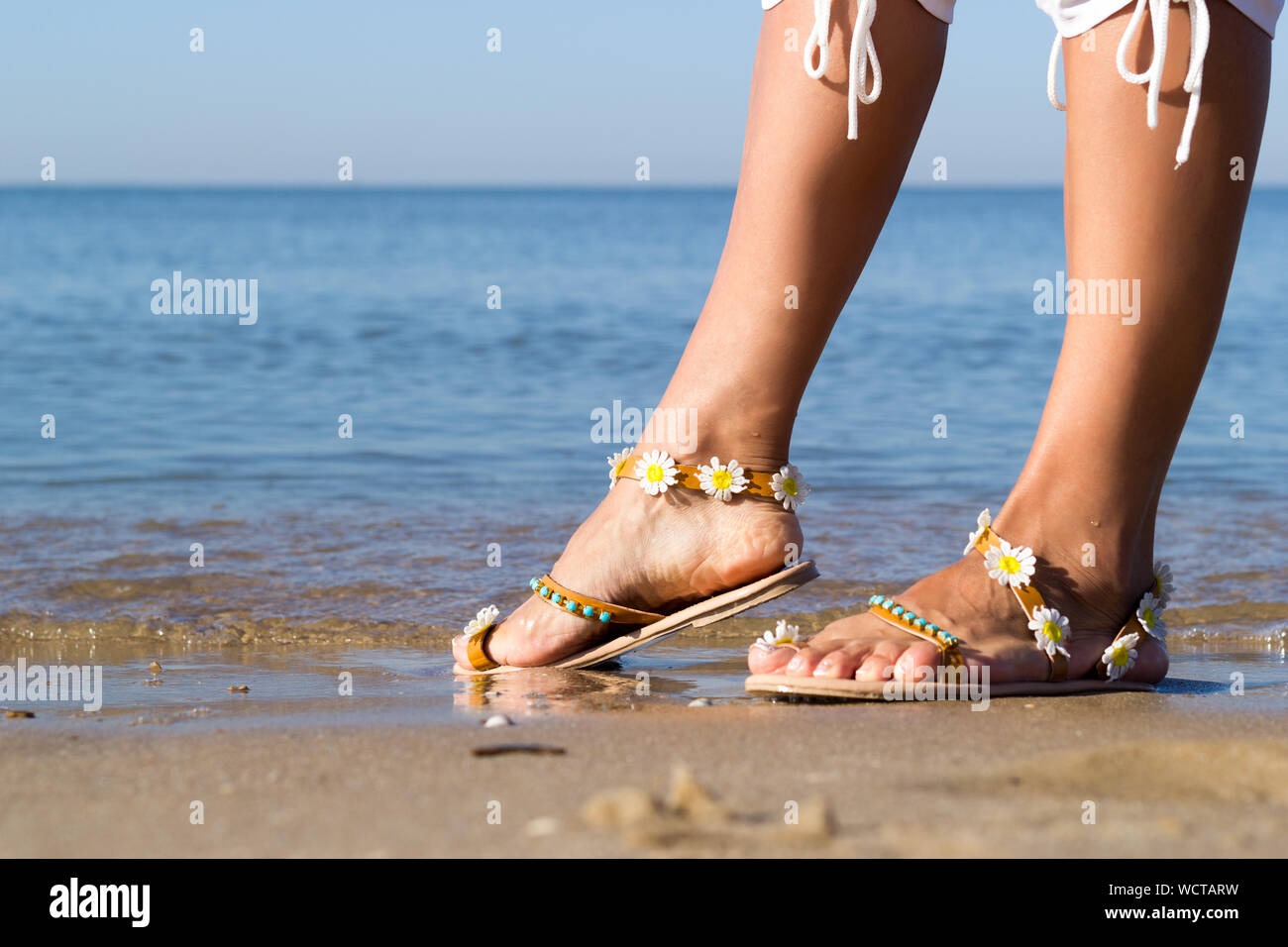 Holidays at sea. Female feet on golden sand on seashore. Woman legs dressed in white shorts and sandals with flowers. Summer relaxation on seacoast. L Stock Photo