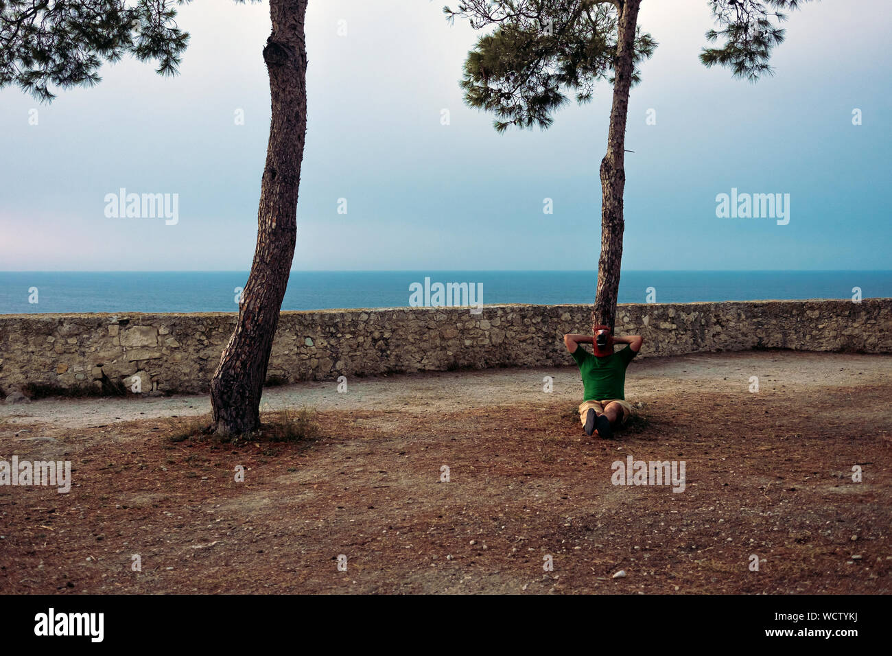 man with a horse head mask relaxes sitting under an Italian stone pine with the sea in the background Stock Photo