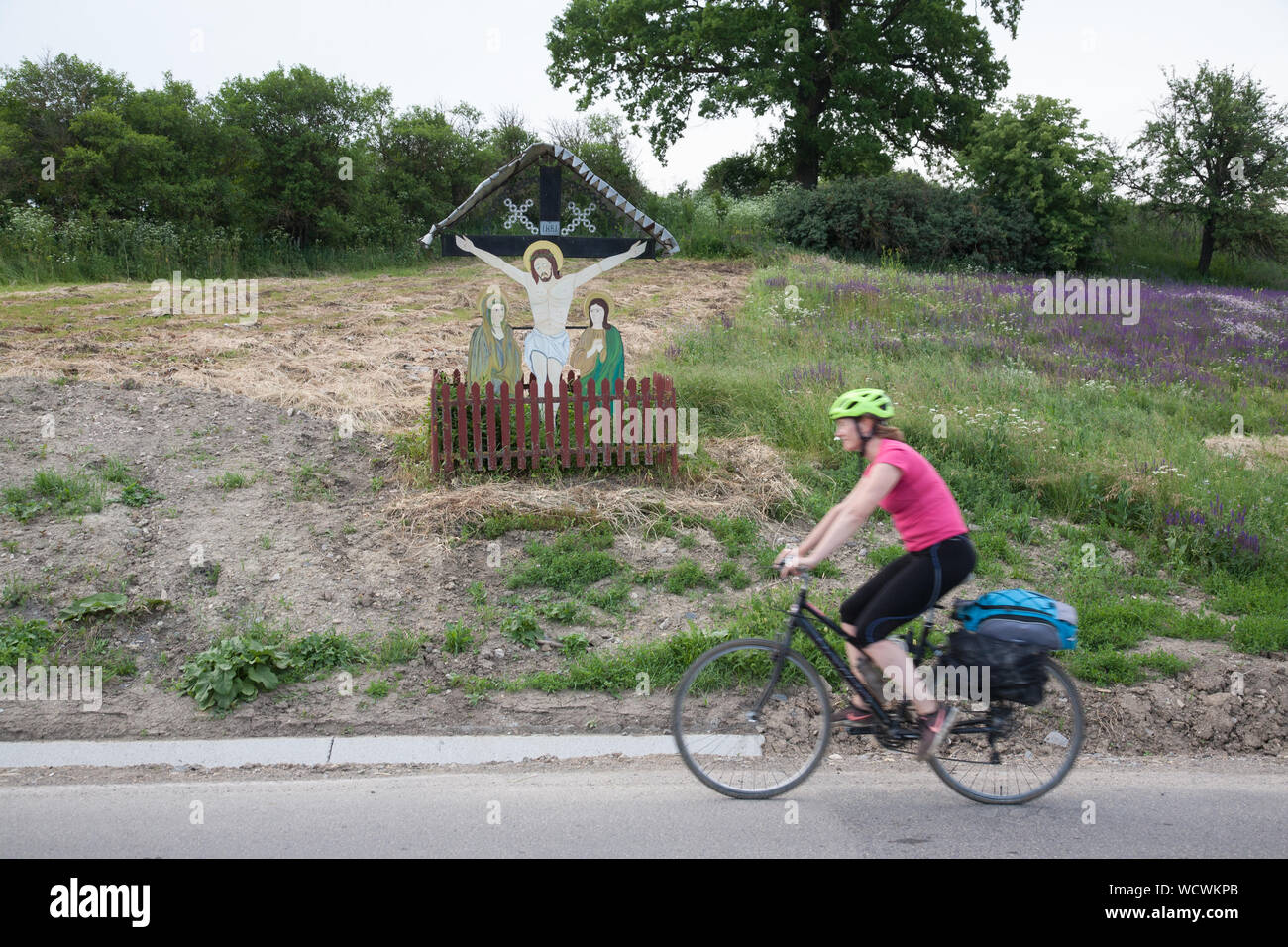 A female cyclist rides past a roadside painted crucifix near the village of Daia, Transylvania, Romania Stock Photo