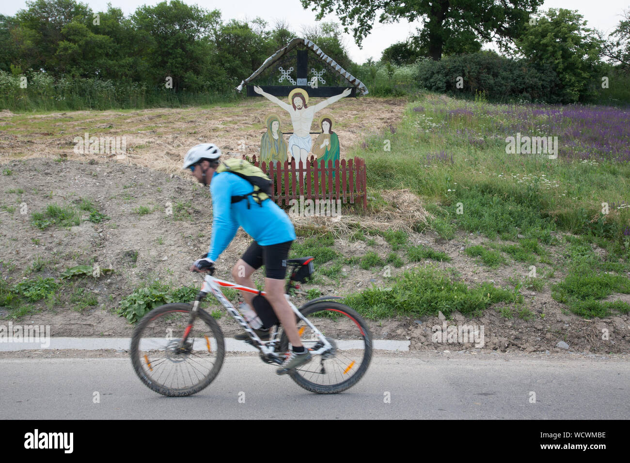A male cyclist rides past a roadside painted crucifix near the village of Daia, Transylvania, Romania Stock Photo