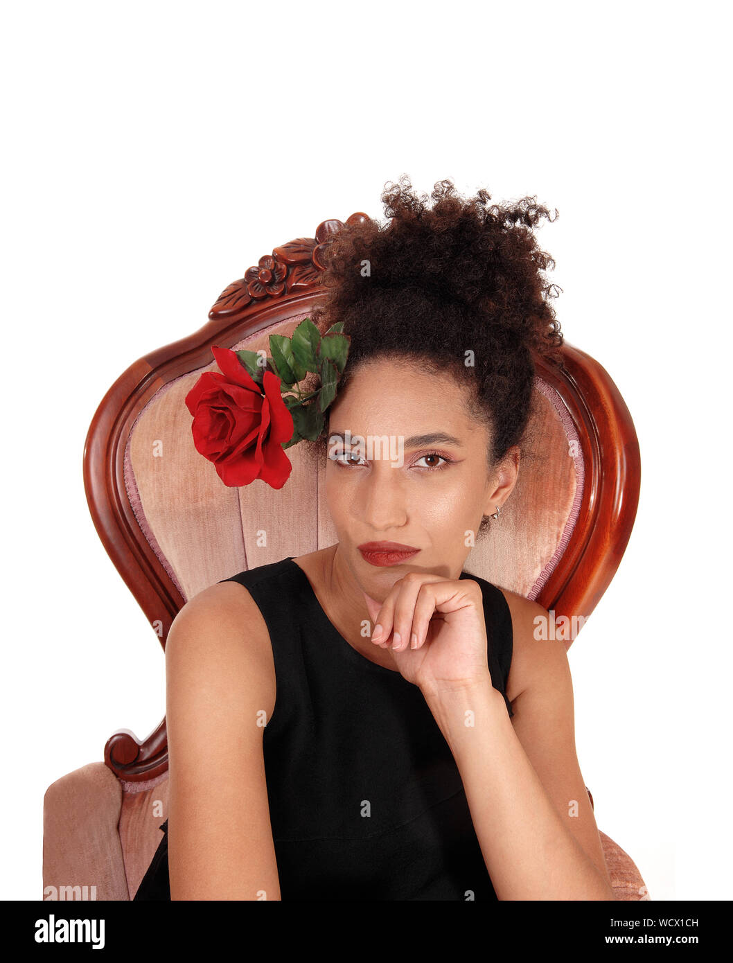 A gorgeous multi-racial woman sitting in a short black dress in an old armchair with a red rose in her hair and her curly black hair in a bun  on her Stock Photo