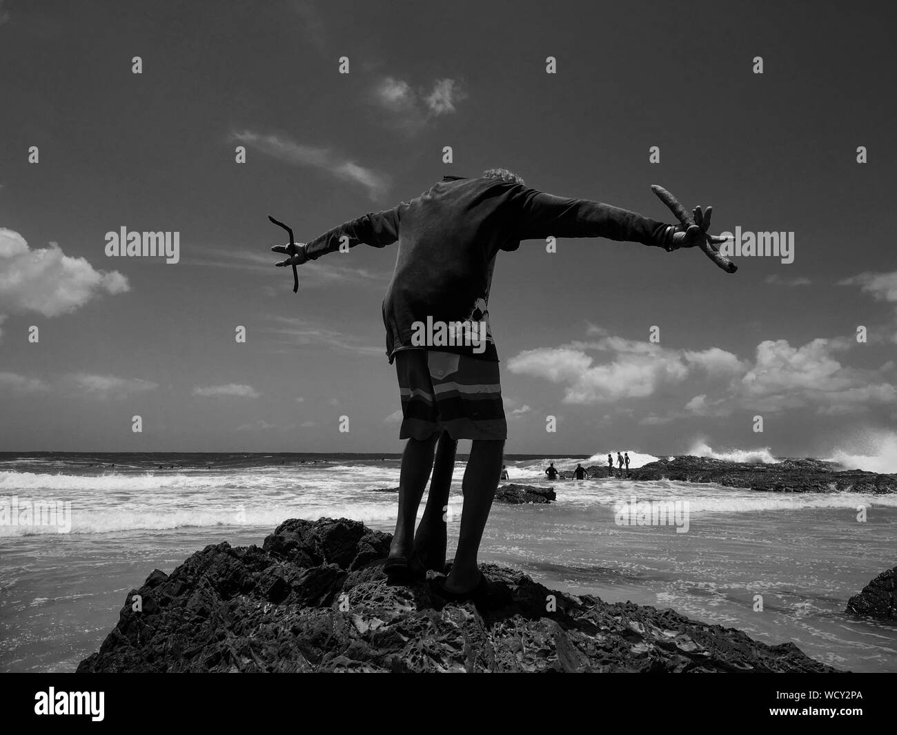 Rear View Of Man Standing With Arms Outstretched On Rock At Rainbow Bay Stock Photo