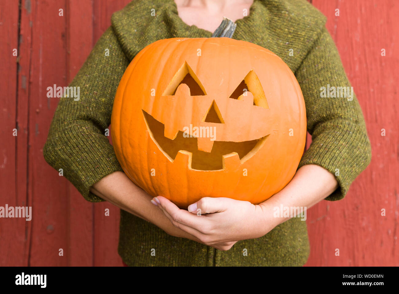 Close-up of young woman holding a pumpkin jack-o'-lantern outdoors in the fall with a red barn in the background Stock Photo