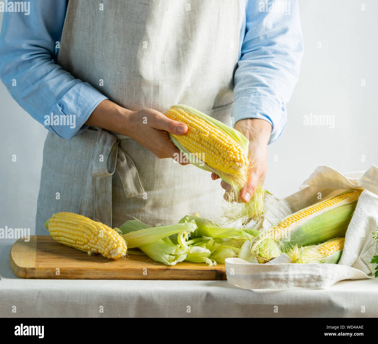 A female cook in a cotton apron cleans ripe corn on the cob. Rustic natural style. The concept of bio farm organic food. Kinfolk style. Copy space Stock Photo