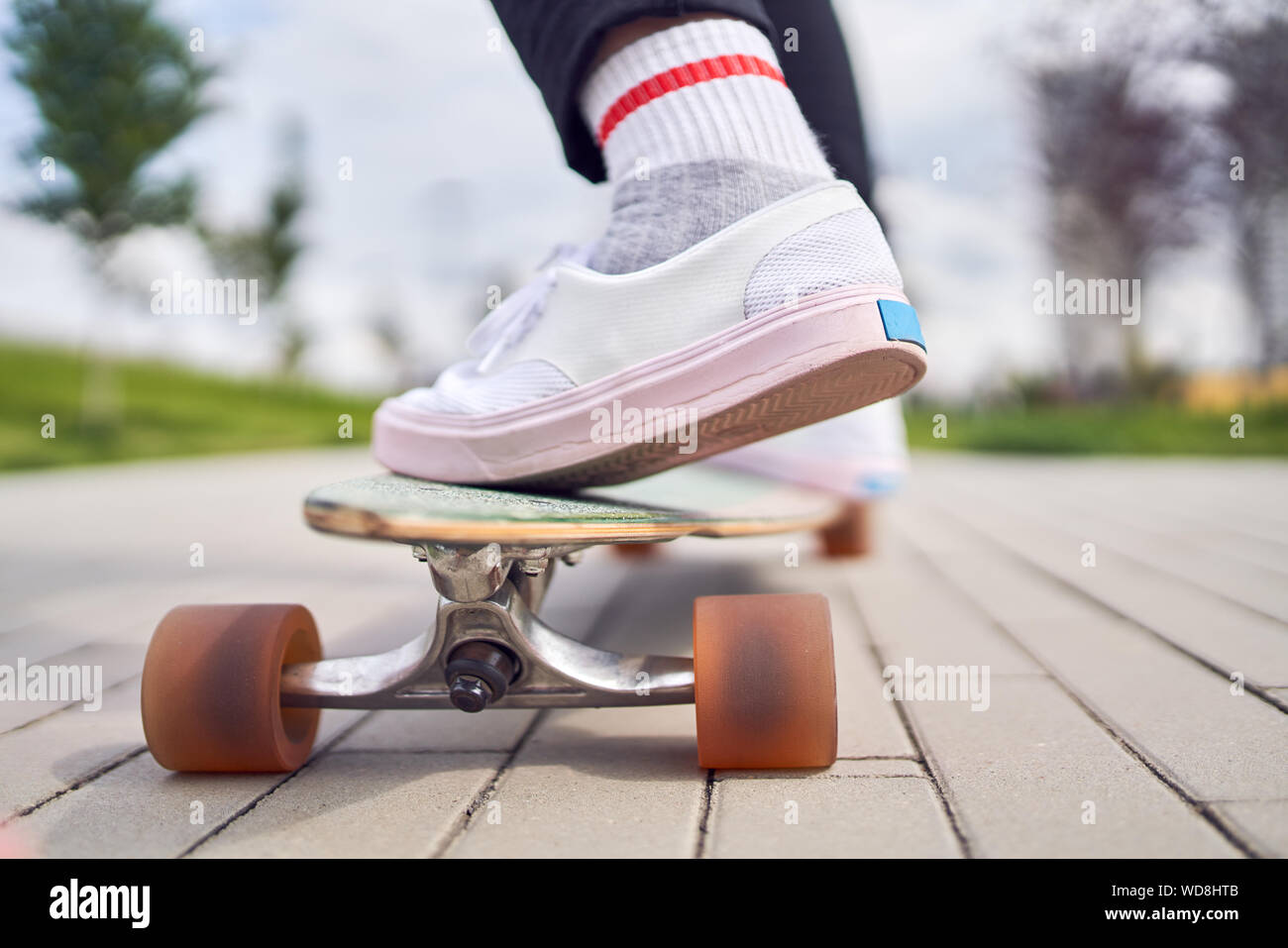 Photo of legs of girl in white shoes riding skateboard on street in city on summer day Stock
