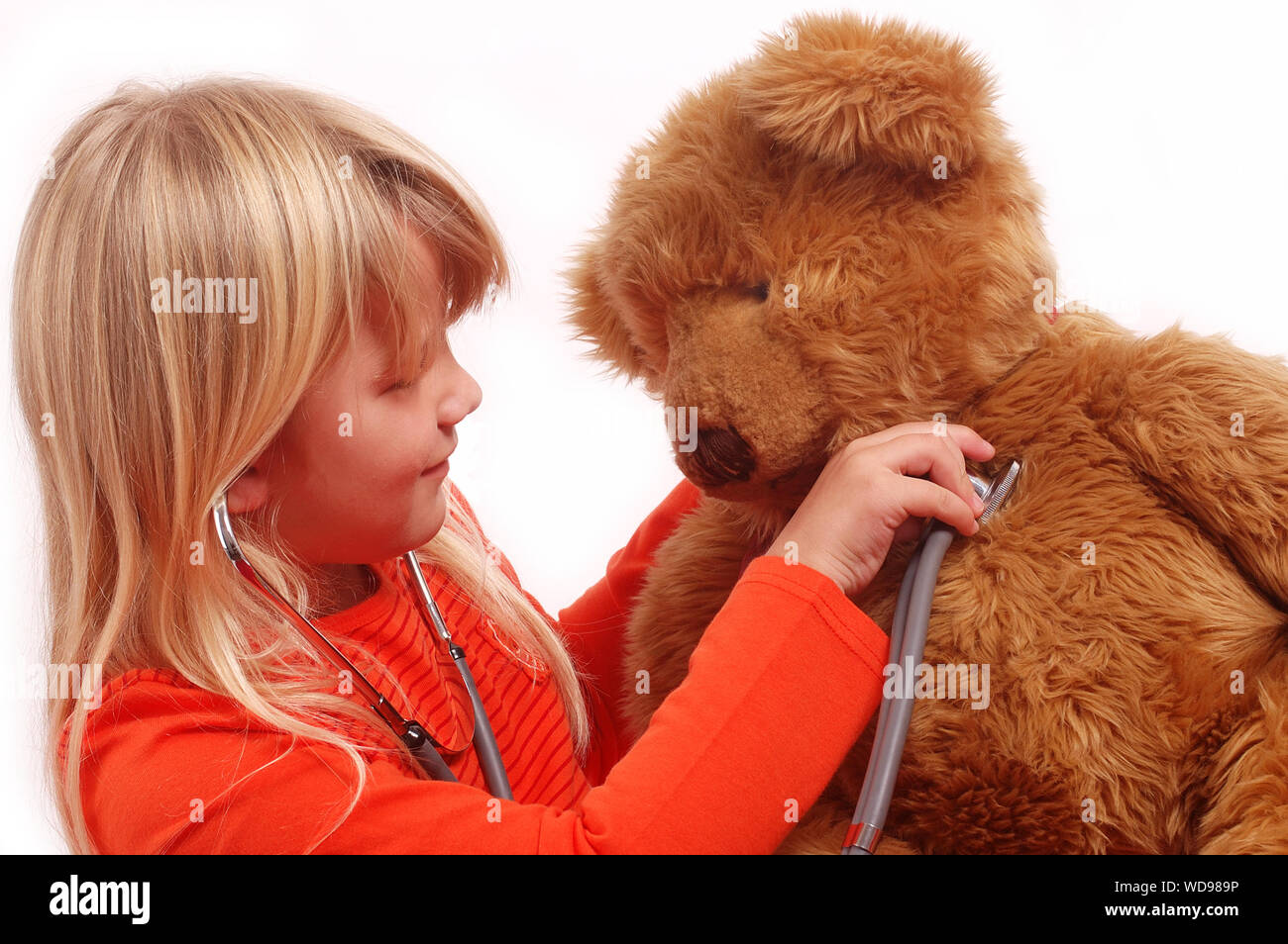 Blonde little girl holding a stethoscope to her big brown teddy bears heart. Stock Photo