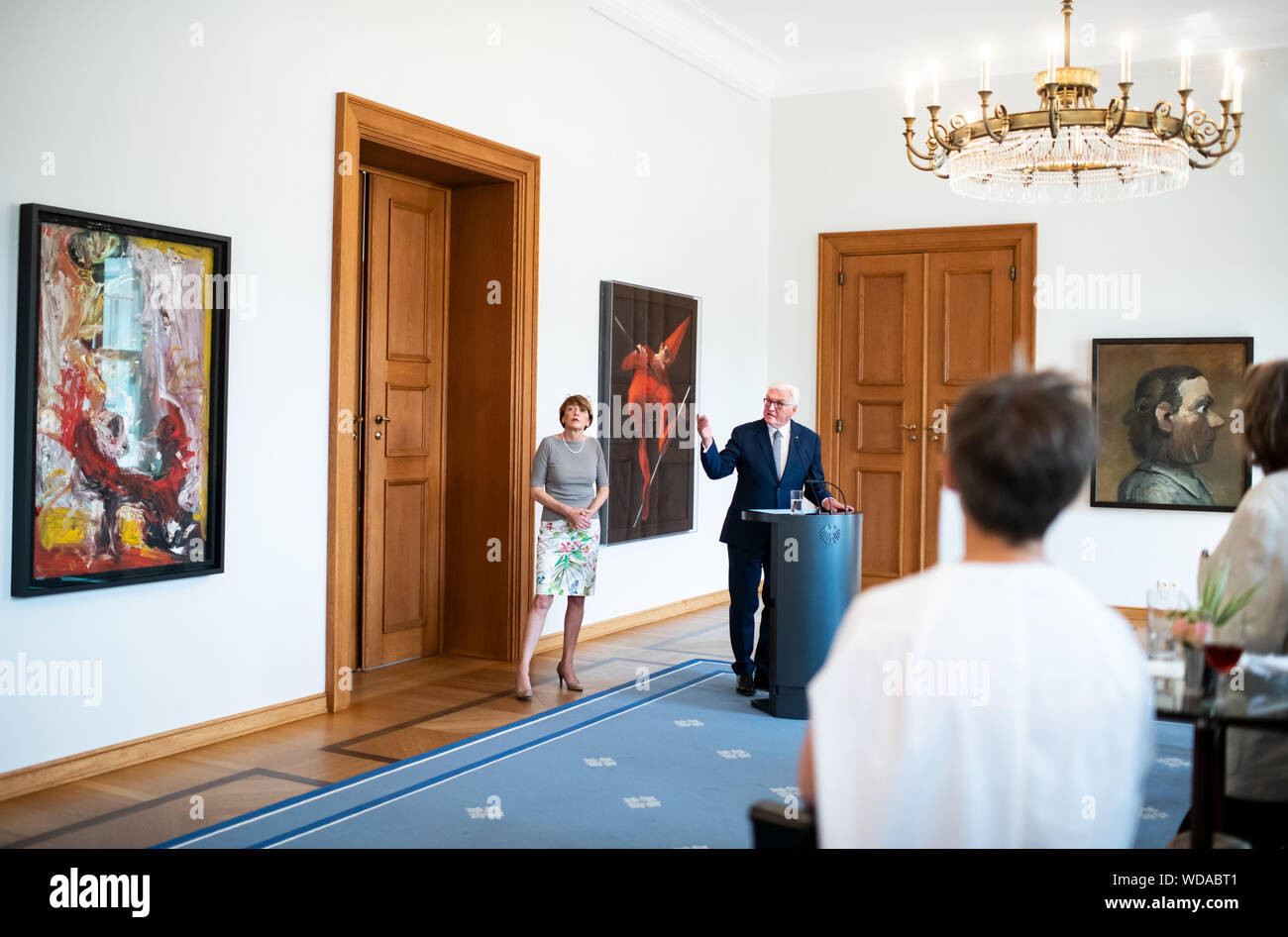 29 August 2019, Berlin: Federal President Frank-Walter Steinmeier speaks in the presence of his wife Elke Büdenbender (l) on the occasion of the presentation of the new equipment of the gallery with paintings from the GDR in Schloss Bellevue. Behind him hangs the painting 'Seiltänzer' (1984) by Trak Wendisch, to the right 'Januskopf' (1977) by Harald Metzkes and to the left 'Kaspar - upside down in ladies boots' (1987) by Hartwig Ebersbach. The exhibition shows pictures of GDR artists in connection with the peaceful revolution 30 years ago. Photo: Bernd von Jutrczenka/dpa Stock Photo