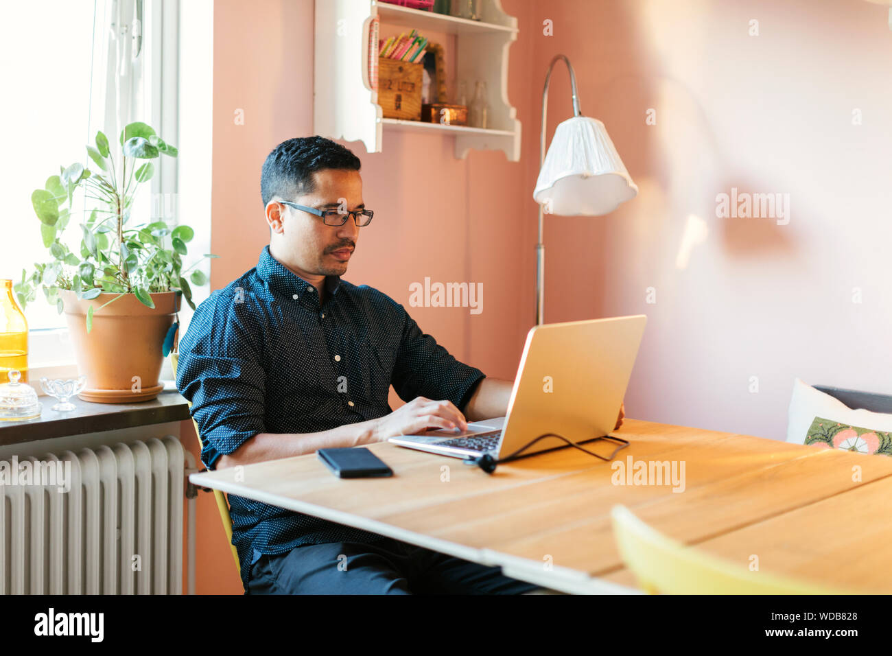 Young man sitting at window and working on laptop. Bearded man sitting at  desktop with smartphone in his hands. Online education. Stock Photo