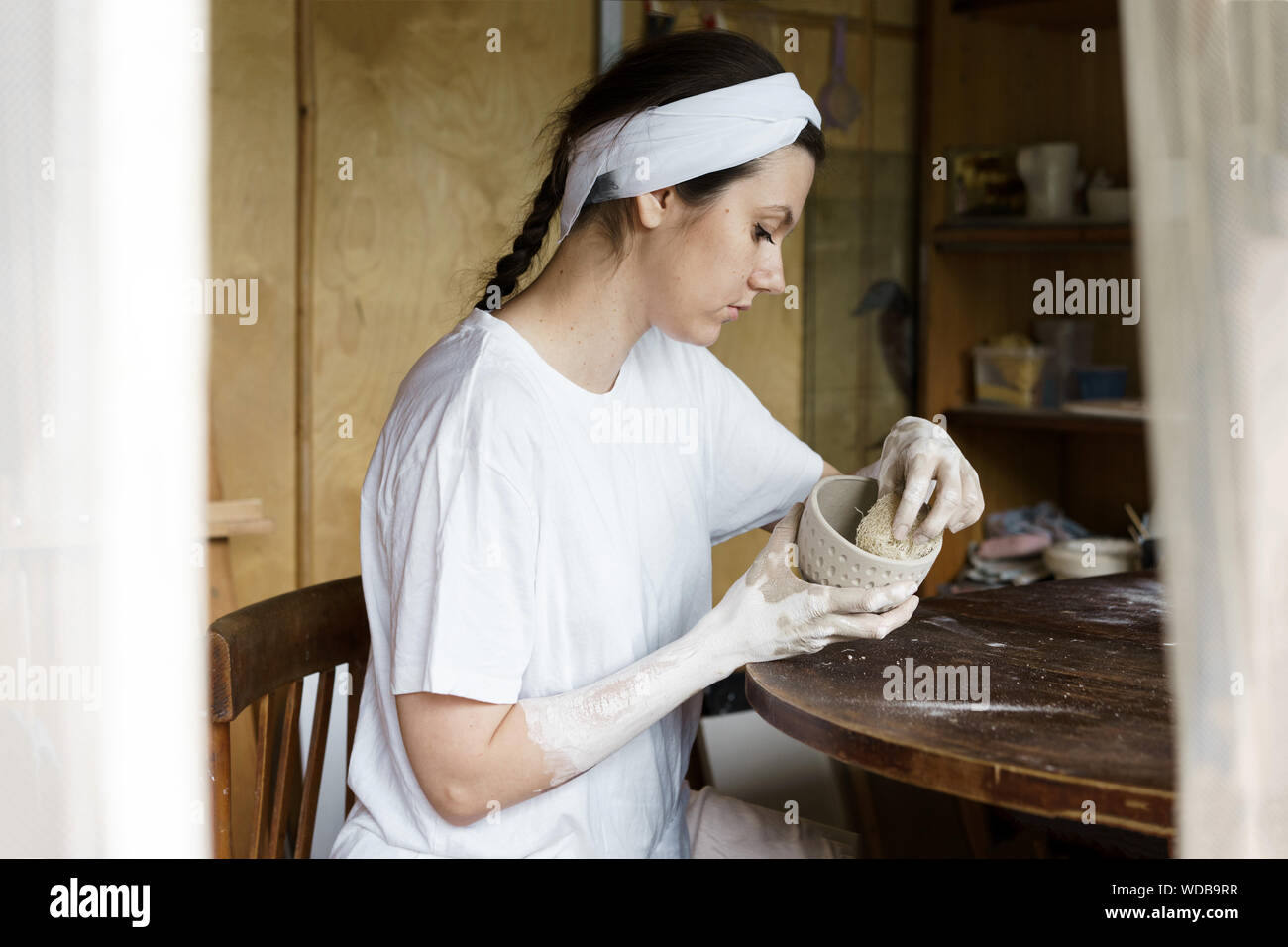 Beautiful woman in a creative workshop sits at a table and wipes a clay bowl Stock Photo
